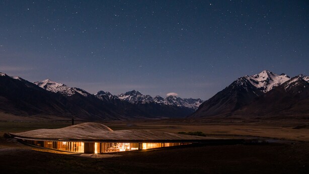 A modern, illuminated building with a curved roof, surrounded by snow-capped mountains under a starry night sky. - The Lindis (Birchwood)
