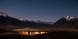 A modern, illuminated building with a curved roof, surrounded by snow-capped mountains under a starry night sky. - The Lindis (Birchwood)
