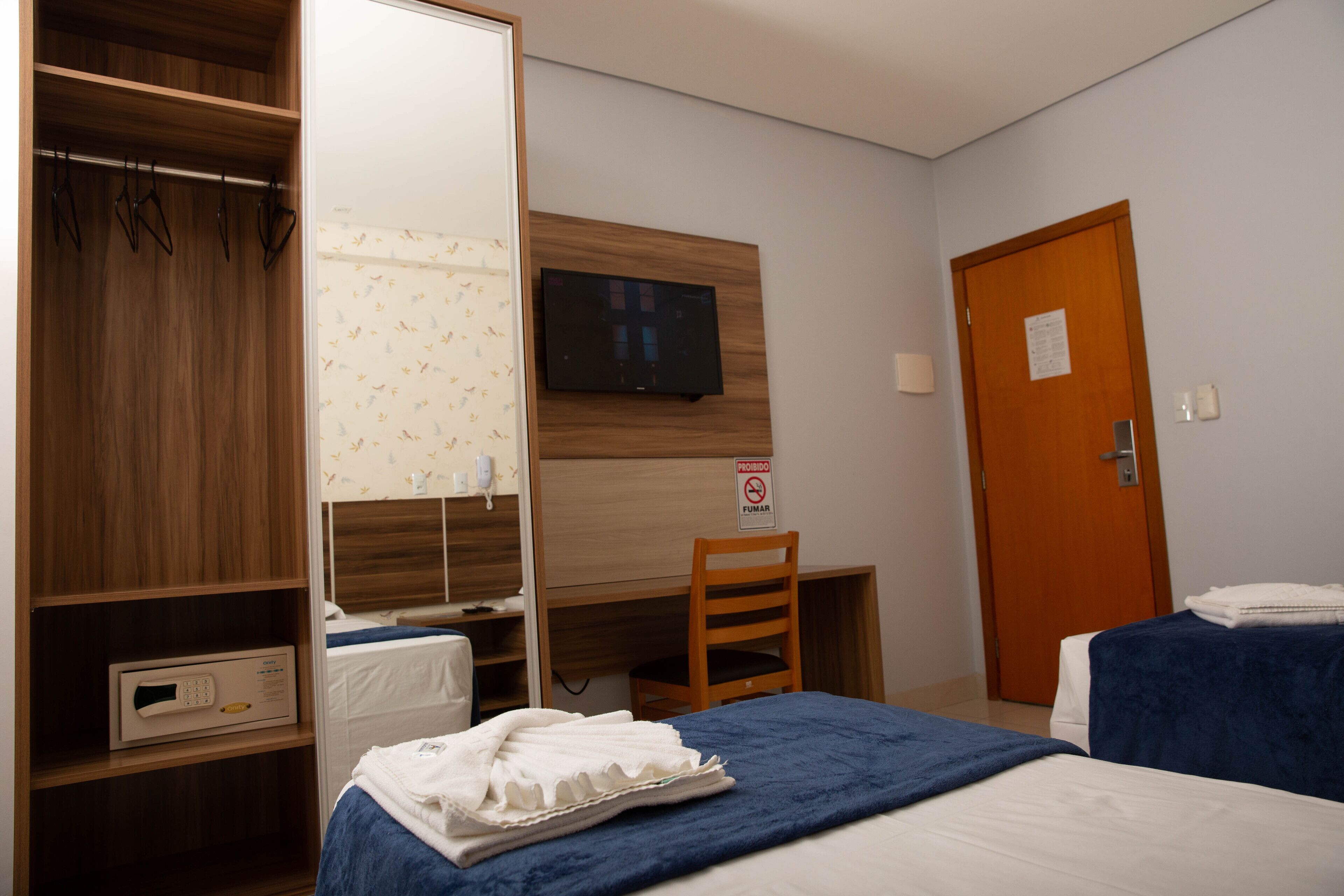 A hotel room with a wooden wardrobe, a desk with a television, a bed with a blue blanket, and a door with a sign.