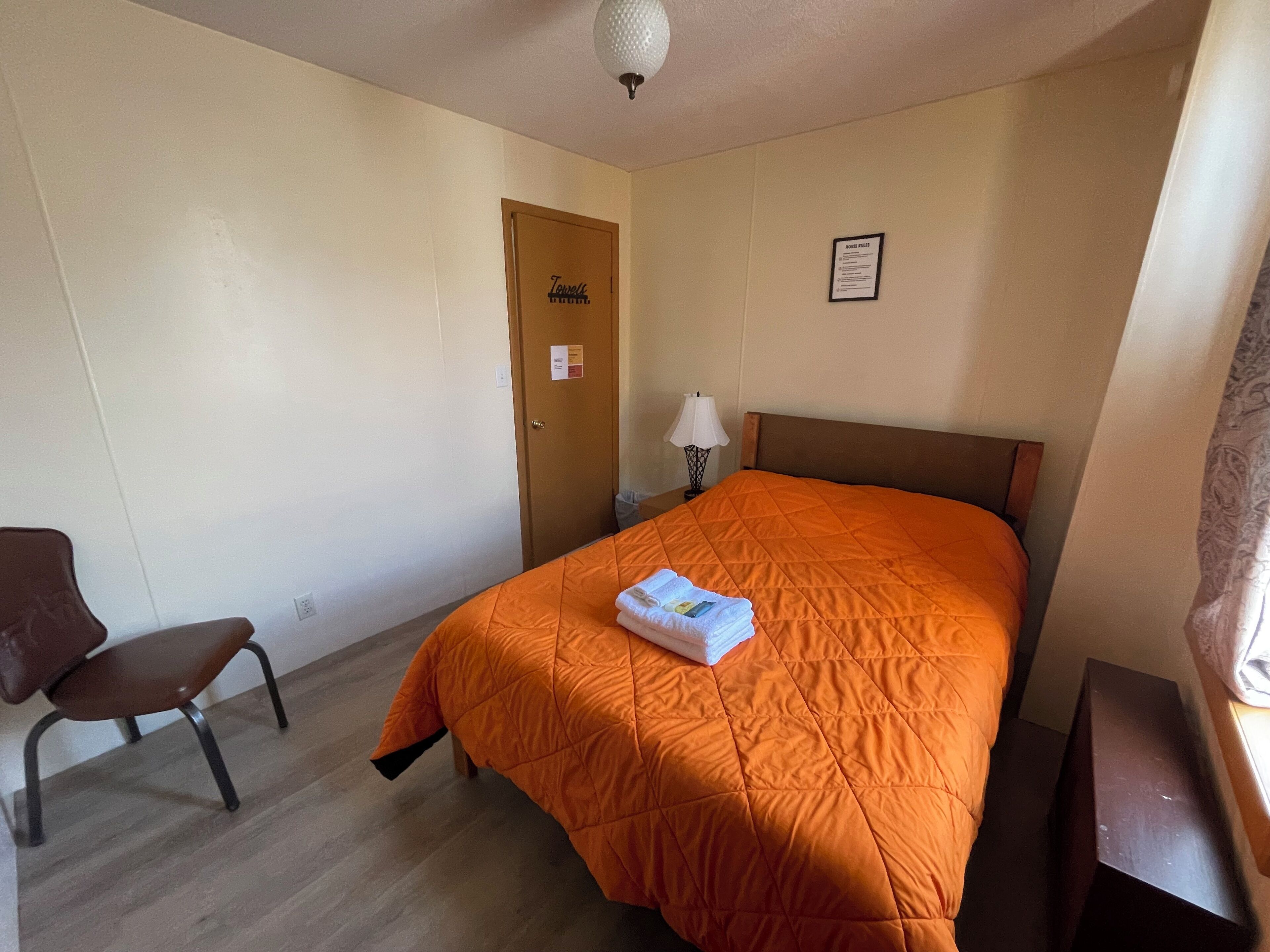 Habitación de hotel con cama, una silla, una mesita de noche con lámpara, una puerta y un cartel en la pared.