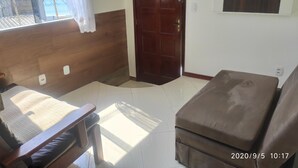Une pièce avec une tête de lit en bois, un canapé marron, une chaise en bois, un rideau blanc et une porte.