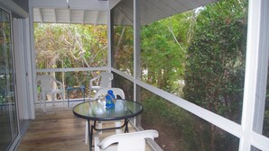 Un espacio al aire libre cubierto, con una mesa de vidrio y sillas de plástico blanco, con vista a un jardín con árboles y arbustos.