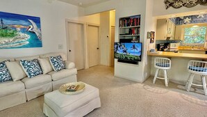 Sala de estar con un sofá color beige, una mesa de centro y un cuadro que representa una escena de playa. Se observa una zona de cocina con una barra y dos taburetes blancos.