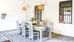 Un coin repas avec une table blanche, des chaises grises et des coussins orange. On y trouve des lampes en osier suspendues, un vase avec des fleurs et une fenêtre ornée d’une grille en métal.