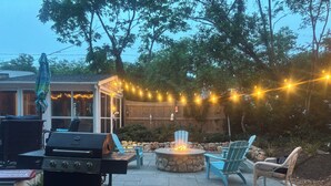 A backyard patio with a stone fire pit, outdoor seating, a grill, and string lights. - Steps To Bay at Campground Beach, A/C, Heat,Wifi, Outdoor Shower, Gas Grill (Eastham)