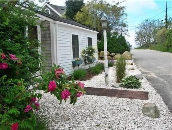 Une petite maison à un seul étage, à l’extérieur blanc et aux volets sombres, entourée d’un jardin où poussent des fleurs roses et bordée d’un chemin de gravier.