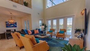 Ein geräumiges Wohnzimmer mit hoher Decke, einem großen Fenster und Blick ins Freie. Mehrere Sitzbereiche mit orange- und blauen Kissen.