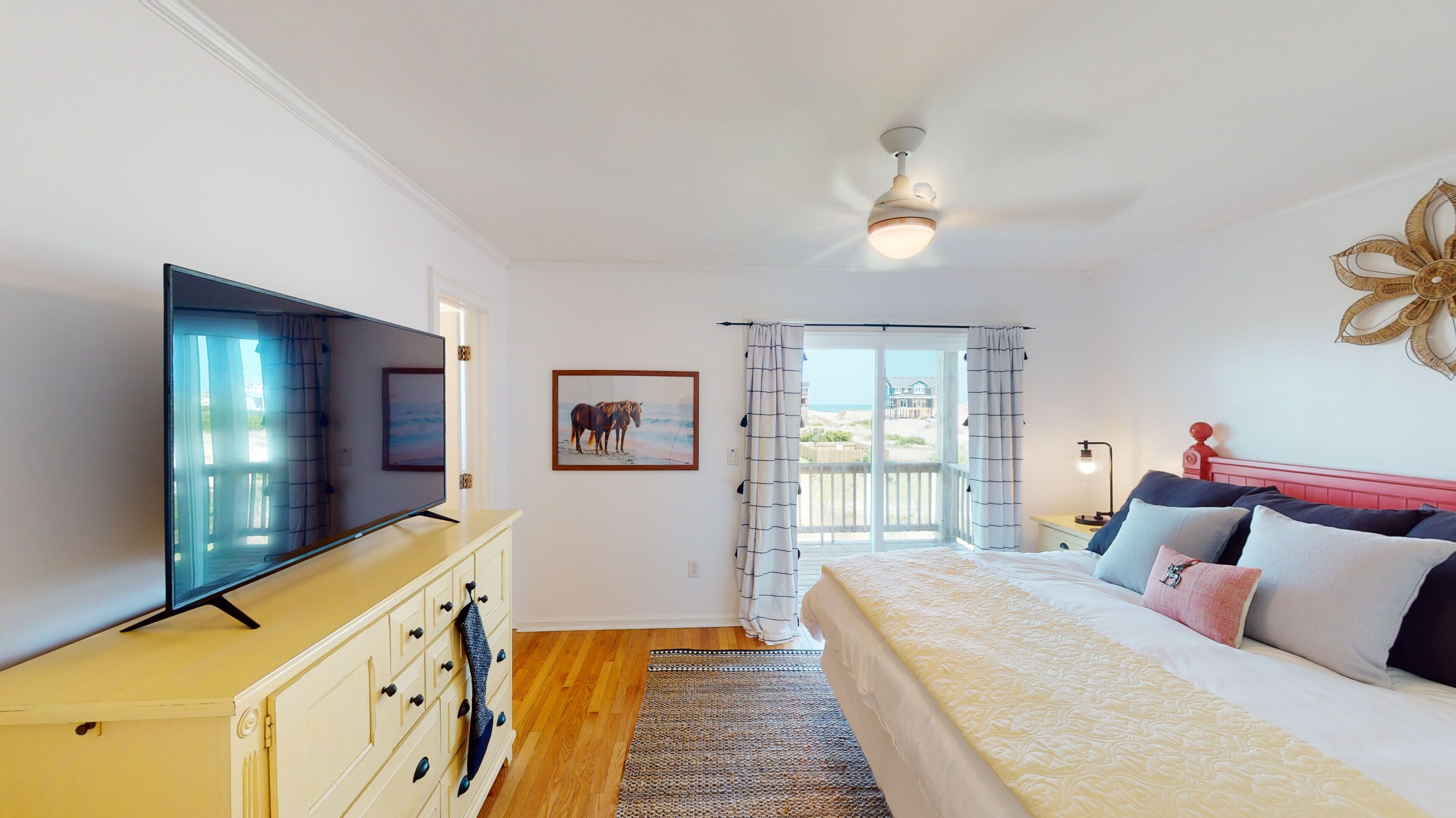 A bedroom with a bed, a TV, a dresser, a window with curtains, and a ceiling fan.