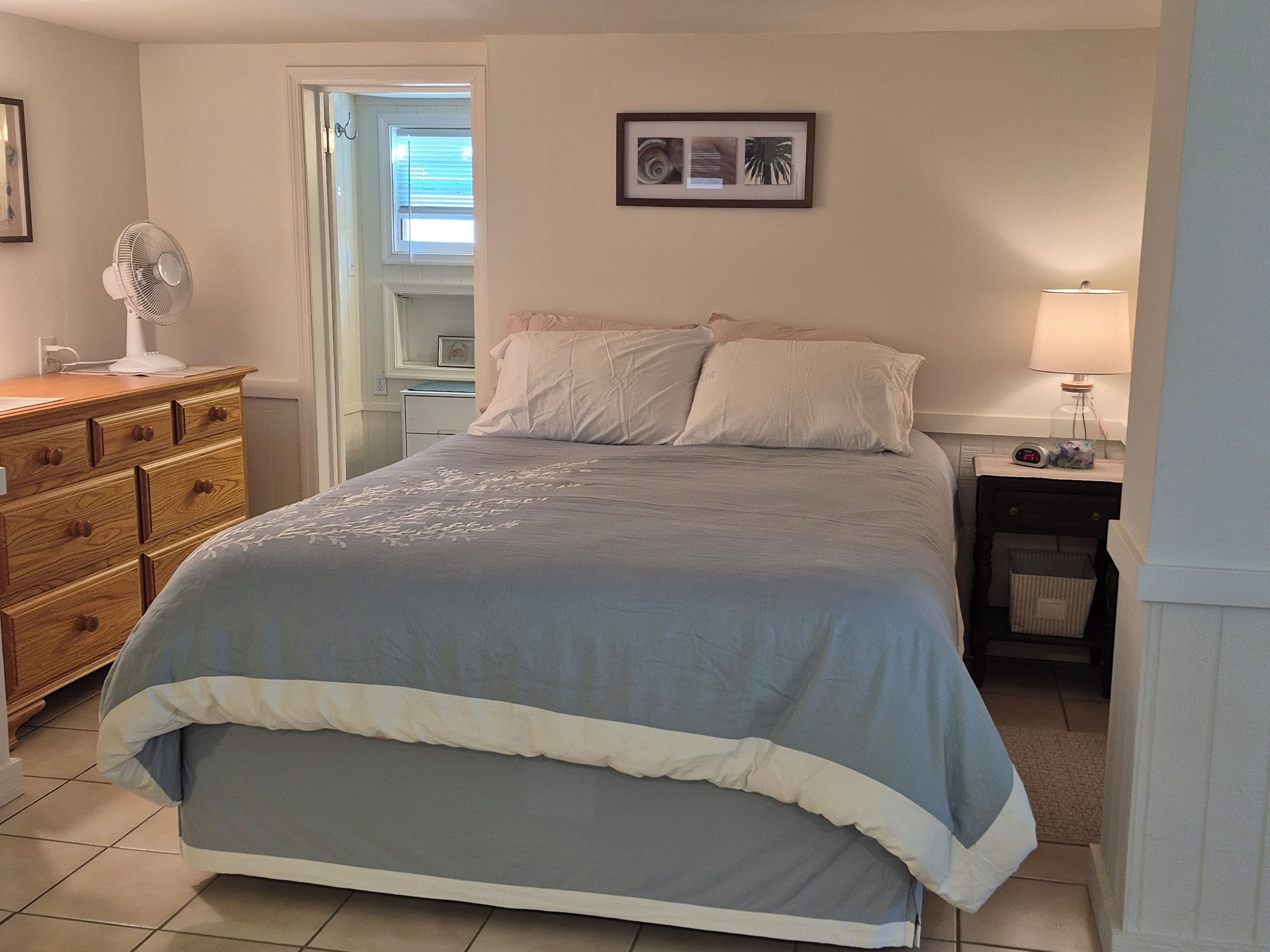 Een slaapkamer met een bed, een dressoir, nachtkastje, lamp en een lijst aan de muur.