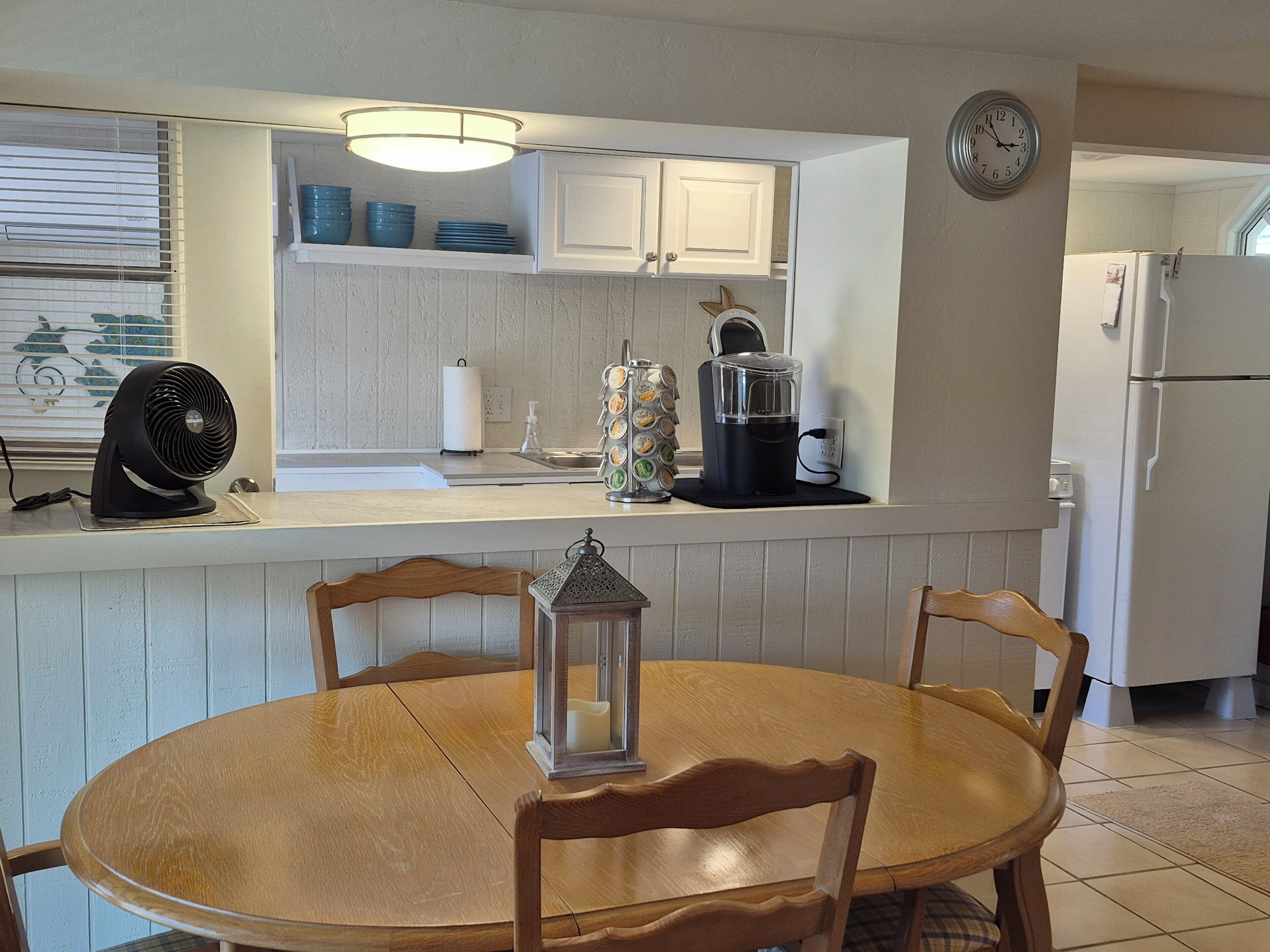 A kitchen with white cabinets, a sink, a stove, a refrigerator, a clock, and a dining table with chairs.