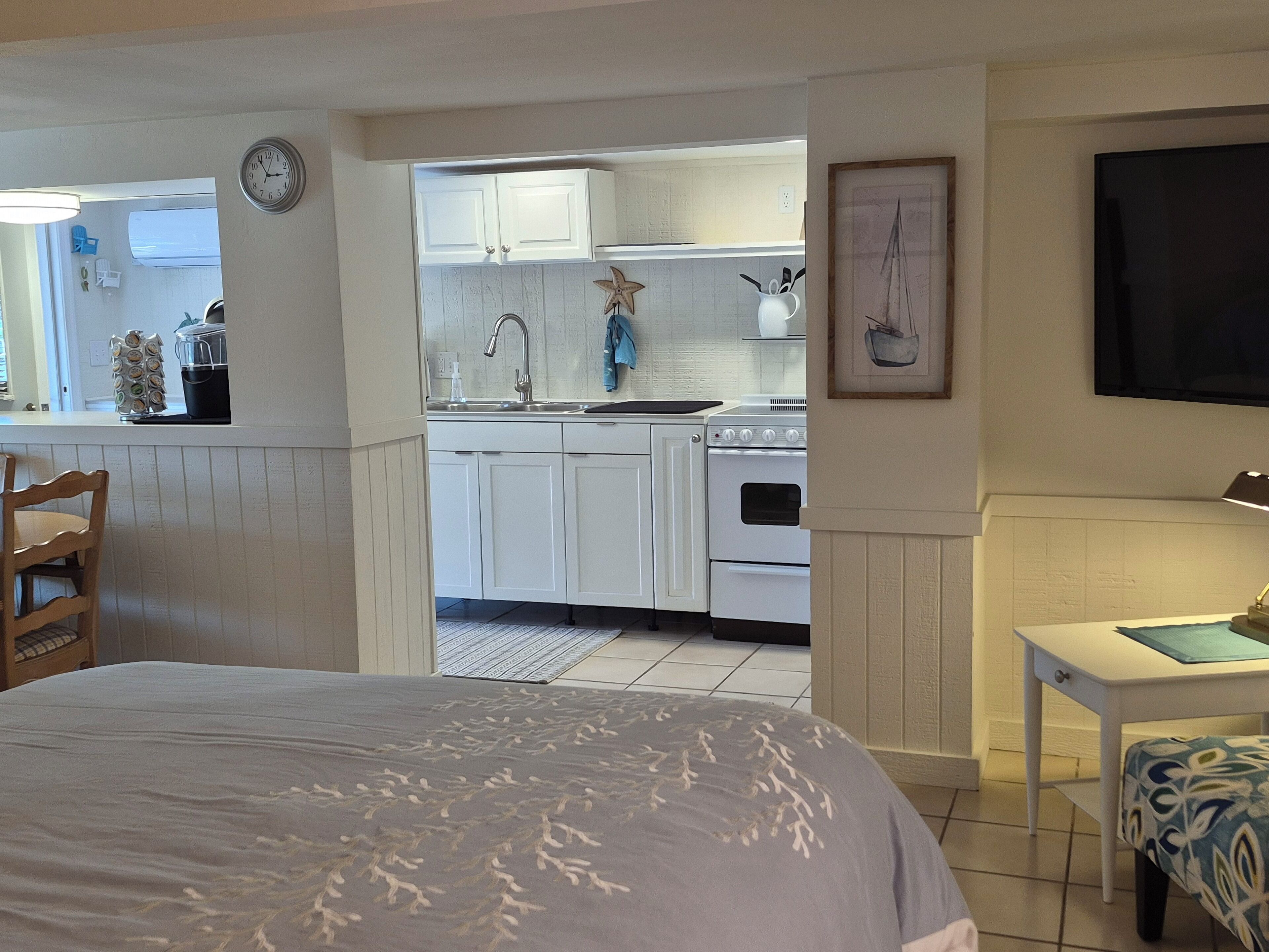 Un dormitorio con cama, una mesita de noche con lámpara y vistas a una cocina con armarios blancos y estufa.