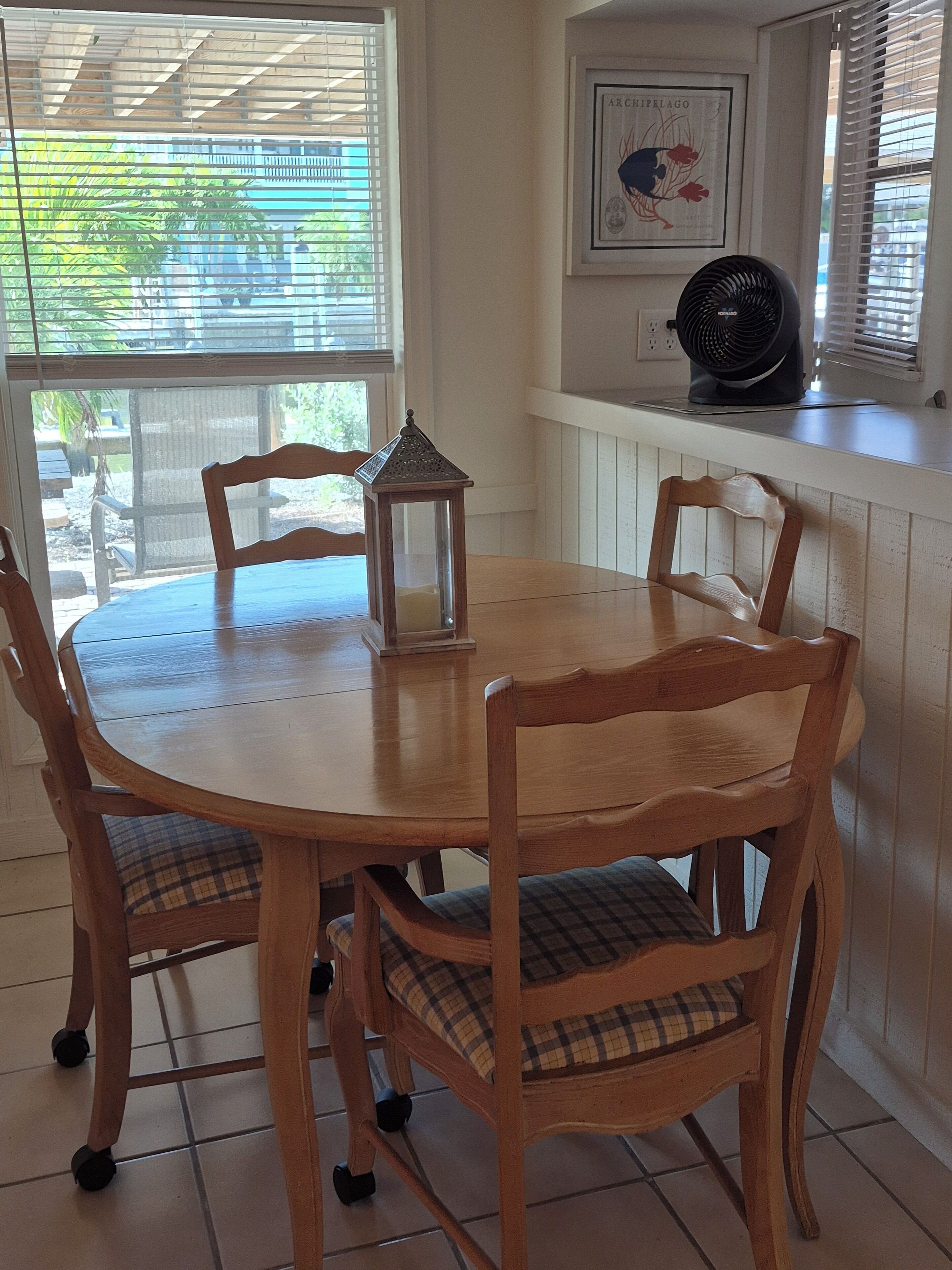 A round wooden dining table with four chairs, a small lantern, and a wall-mounted fan.