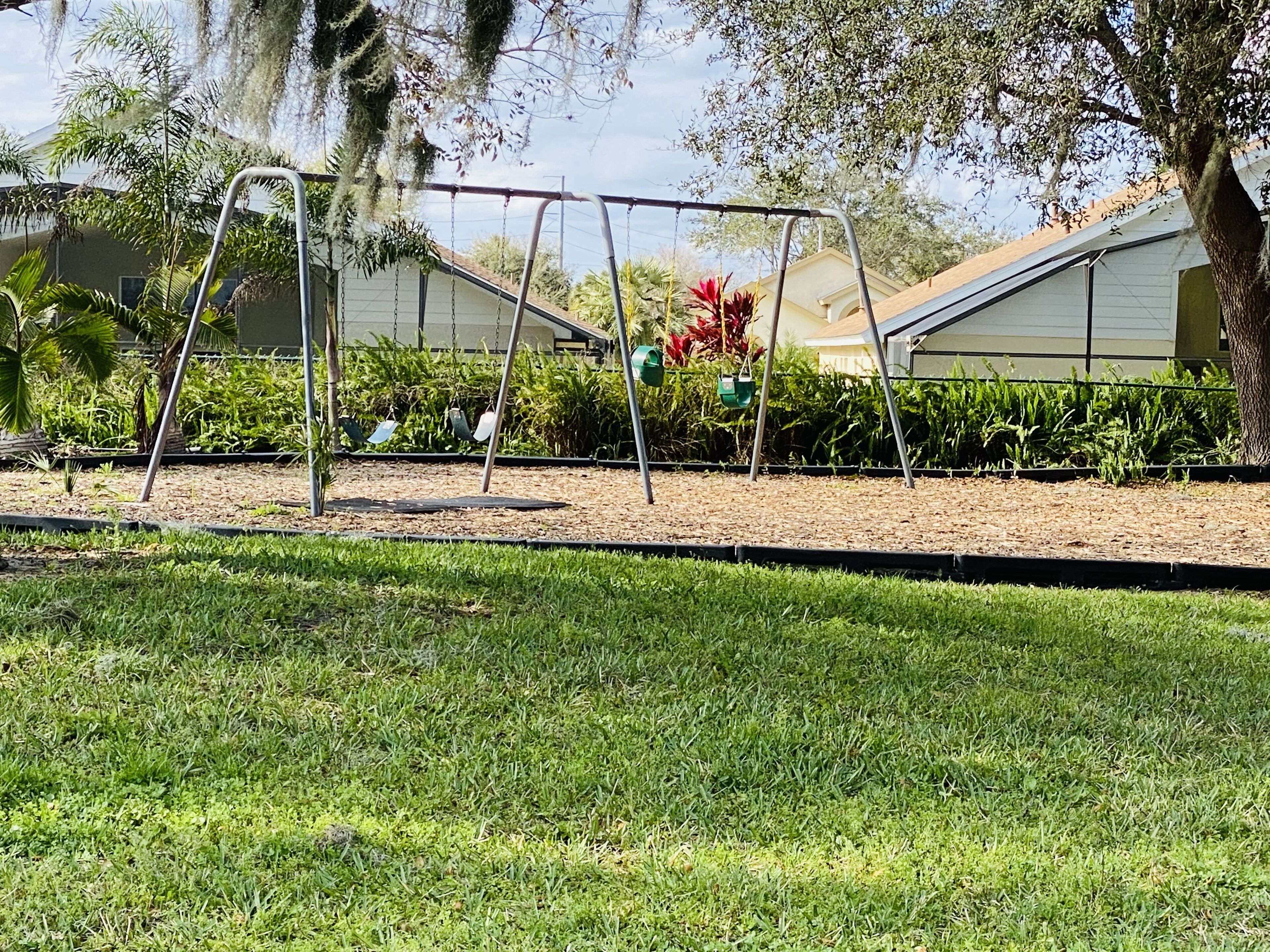 A playground with swings and a sandbox in a grassy area with trees and a building in the background.