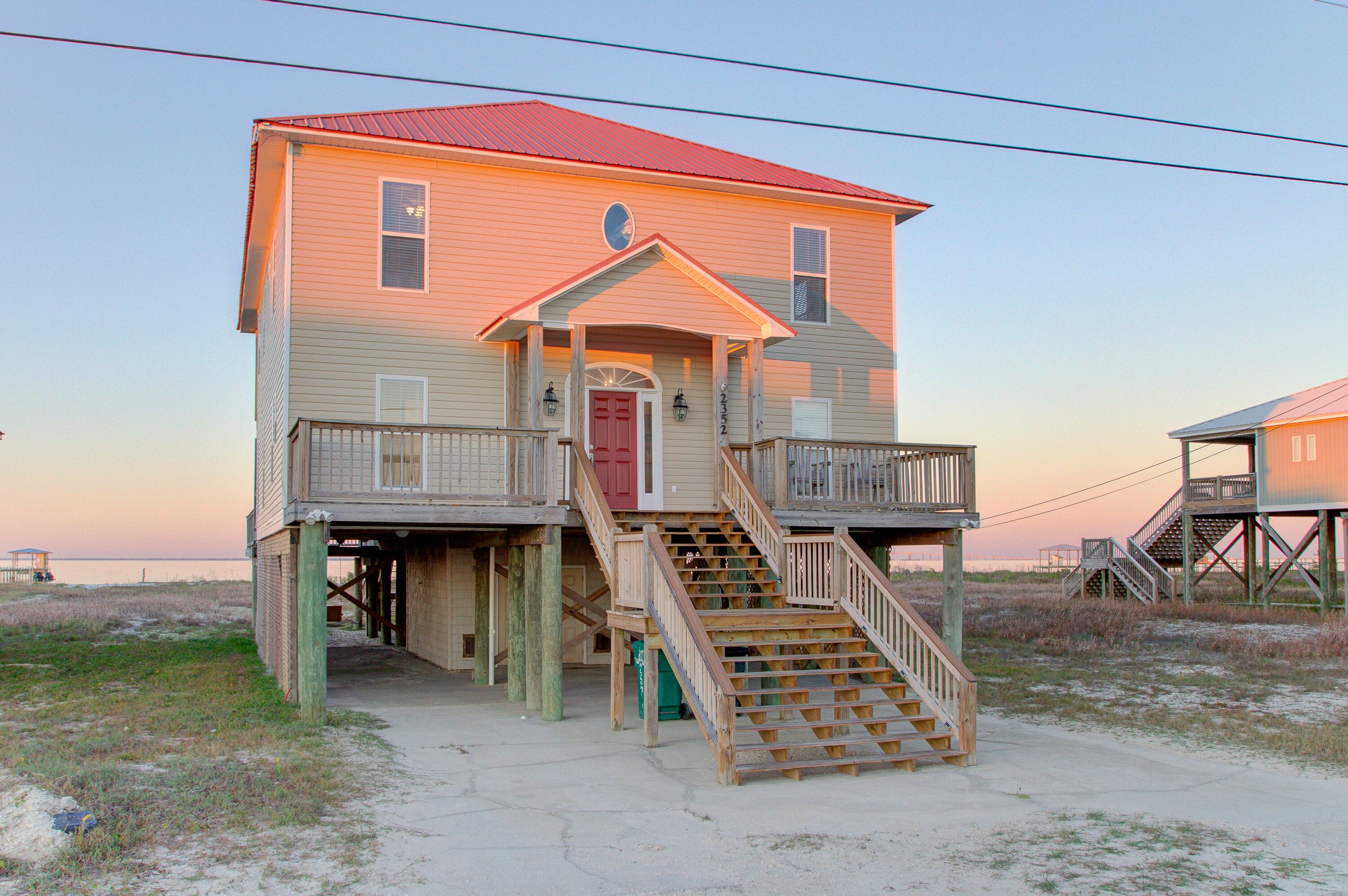 Une maison de plage à deux étages, avec un toit rouge, un porche à l’avant et un escalier menant à l’étage supérieur.