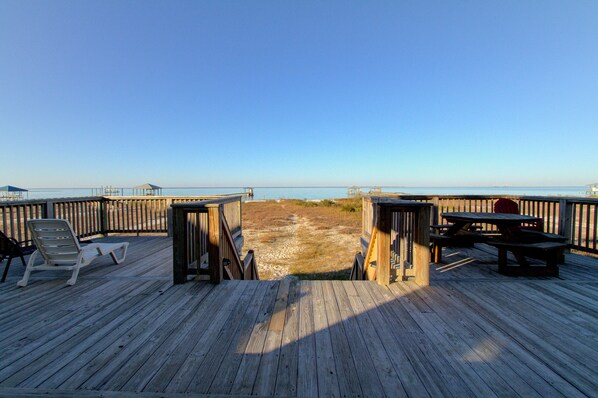 A wooden deck with a view of the beach, a picnic table, and a lounge chair.