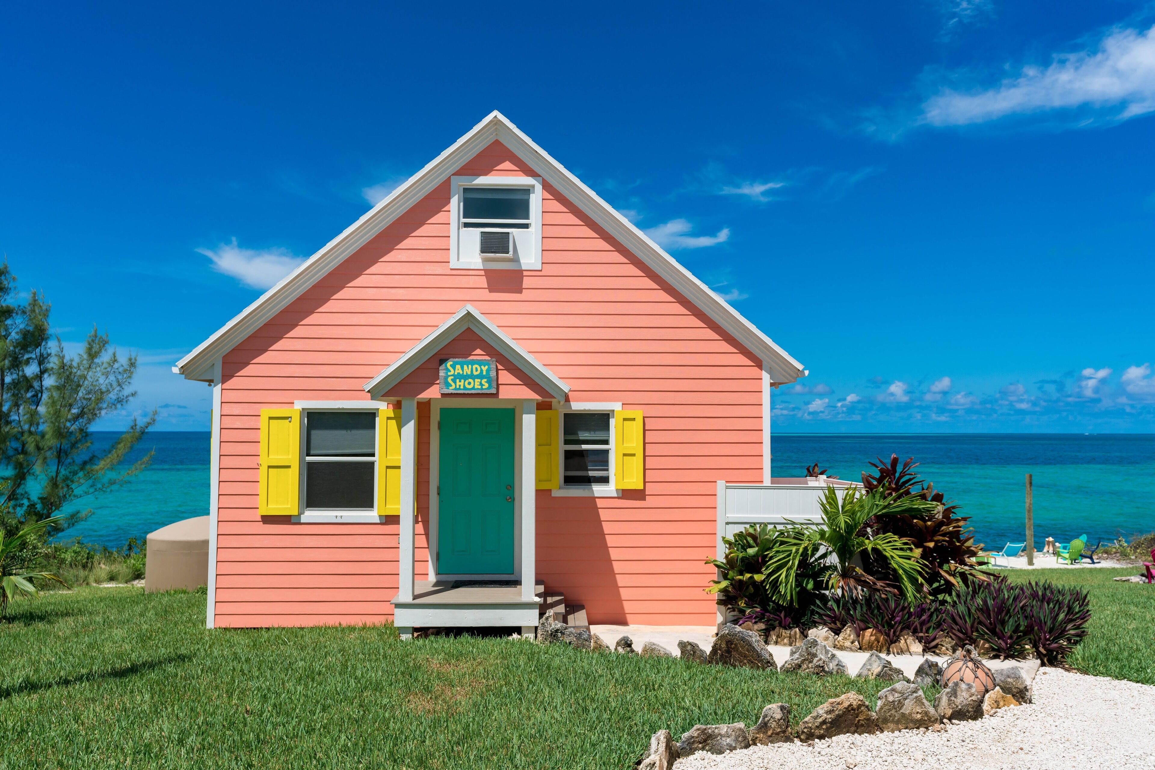 A pink house with a green door and yellow shutters, labeled "Sandy Shoes," situated by a beach with clear blue water and a clear sky.