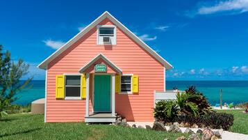 A pink house with a green door and yellow shutters, labeled "Sandy Shoes," situated by a beach with clear blue water and a clear sky.