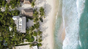 Luftaufnahme eines Anwesens direkt am Strand mit einem Gebäude mit Reetdach, Palmen und einem Sandstrand.