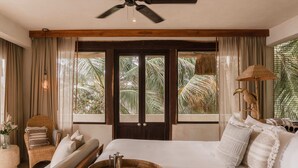 Ein Schlafzimmer mit einem Bett, einem Sessel, einem Fenster mit Vorhängen, einem Deckenventilator und Blick auf Palmen.