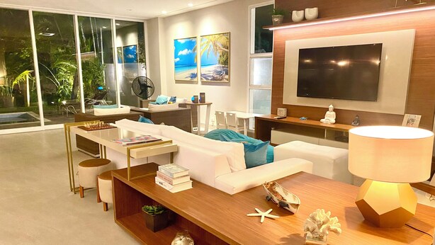 A modern living room with a wooden coffee table, white sofas, a TV mounted on the wall, and a large window with a view of greenery.