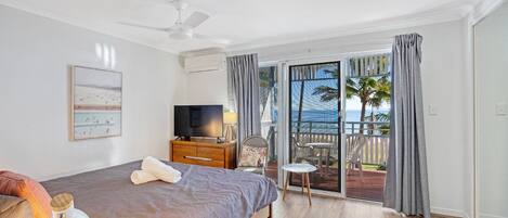Ein Schlafzimmer mit Bett, Fernseher, Deckenventilator, Fenster mit Vorhängen und Blick auf den Strand.