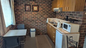 A compact kitchen with a brick wall, wooden cabinets, a small sink, a microwave, and a chair. - Barham Colonial Motel (Barham)