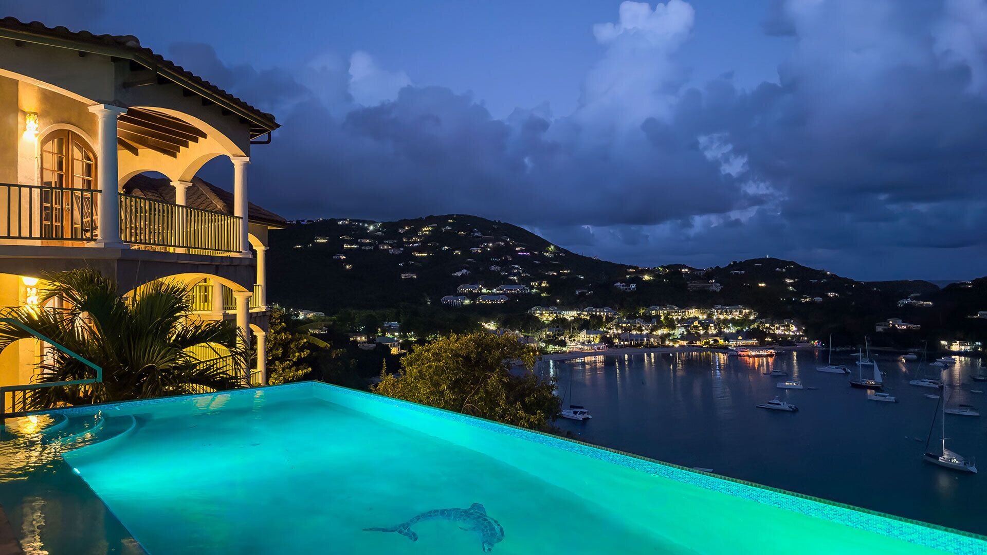 Une piscine avec vue sur la mer, un balcon illuminé et un paysage montagneux en arrière-plan.