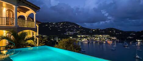Une piscine avec vue sur la mer, un balcon illuminé et un paysage montagneux en arrière-plan.