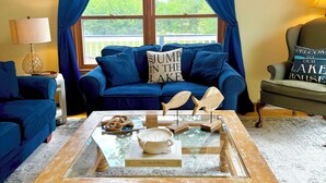 Uma sala de estar com um sofá azul, uma mesa de centro com objetos decorativos, uma janela com cortinas azuis e uma placa na parede.