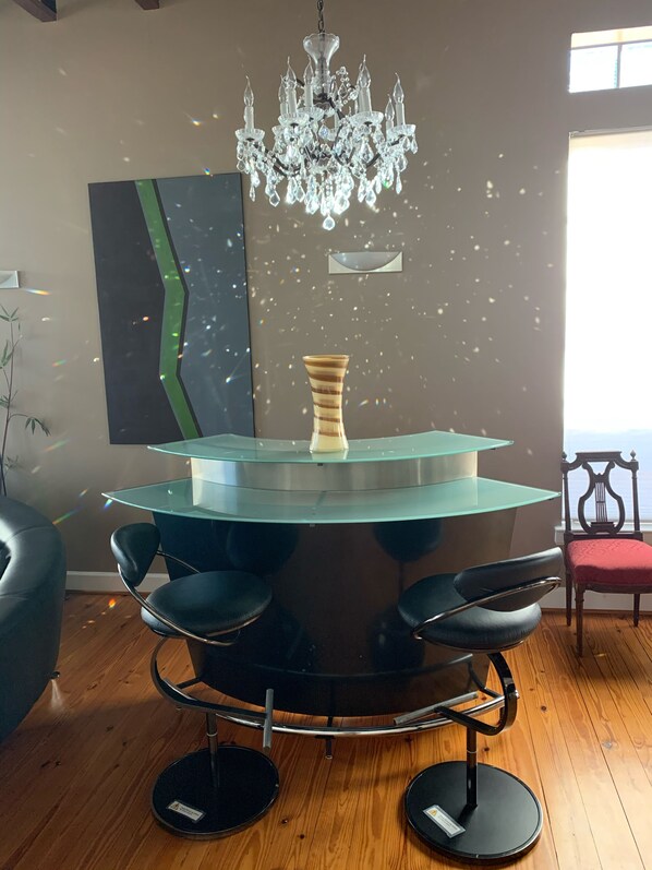 A modern interior with a glass-top bar, black bar stools, a crystal chandelier, a vase, and a large abstract painting.