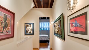 A hallway with framed artworks on the walls, a bed in the background, and a wooden floor. - Montrose NY Style Brick Home with a chef kitchen! (Houston)