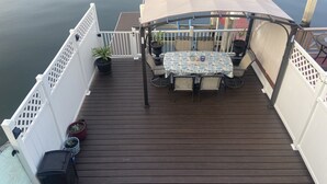 A wooden deck with a table and chairs, surrounded by white railings, overlooking a calm body of water.