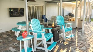 Ein überdachter Patio-Bereich mit türkisfarbenen Adirondack-Stühlen, einer kleinen Feuerstelle und einem an der Wand befestigten Fernseher.