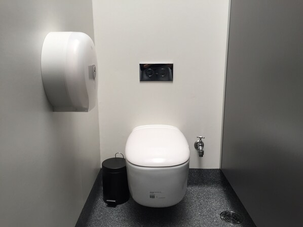 Un baño pequeño con inodoro blanco, un cubo de basura negro y un secador de manos montado en la pared.