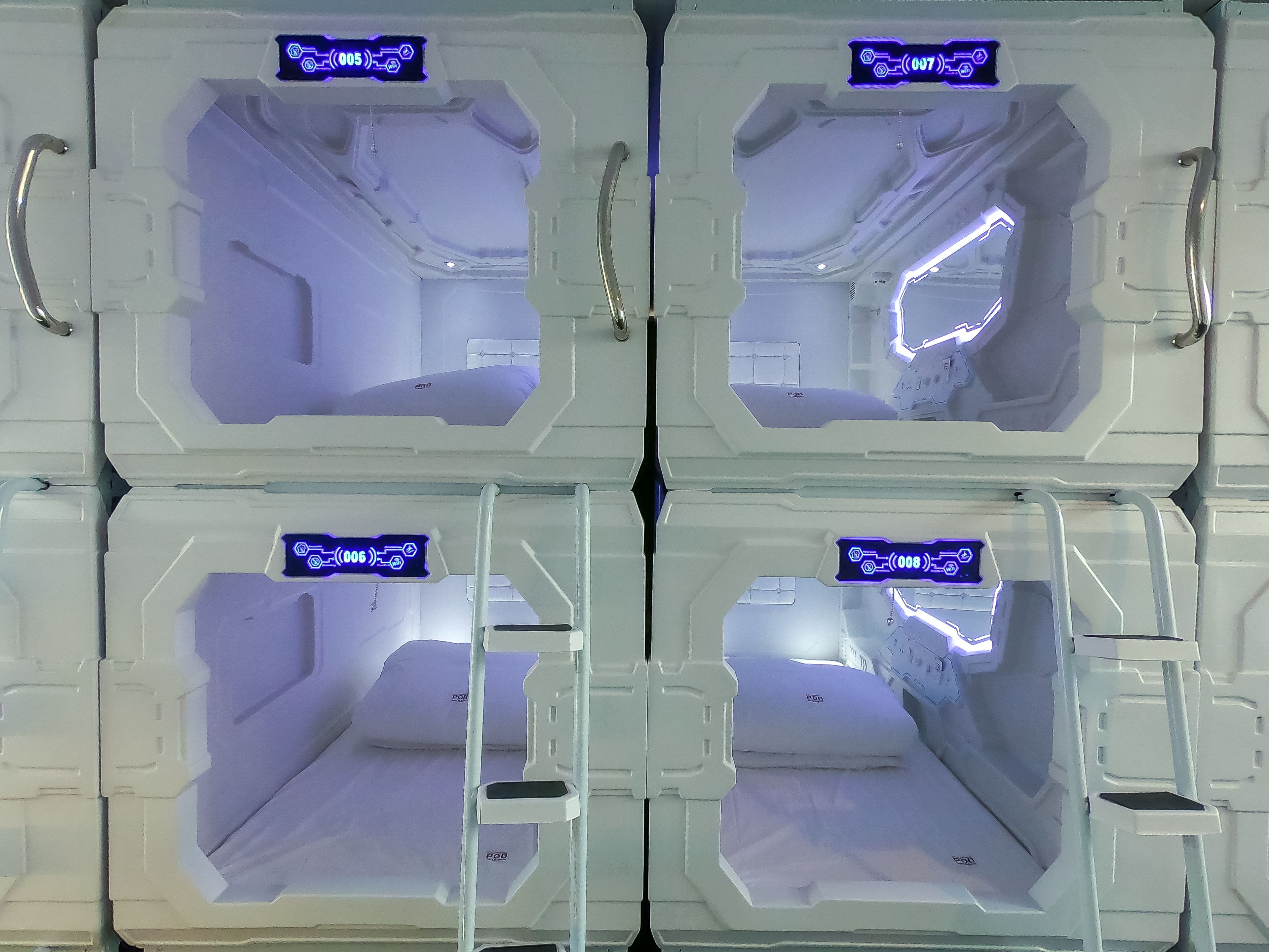 Quatro quartos idênticos, vazios, brancos e com design futurista, com puxadores metálicos e placas iluminadas em azul mostrando os números dos quartos.