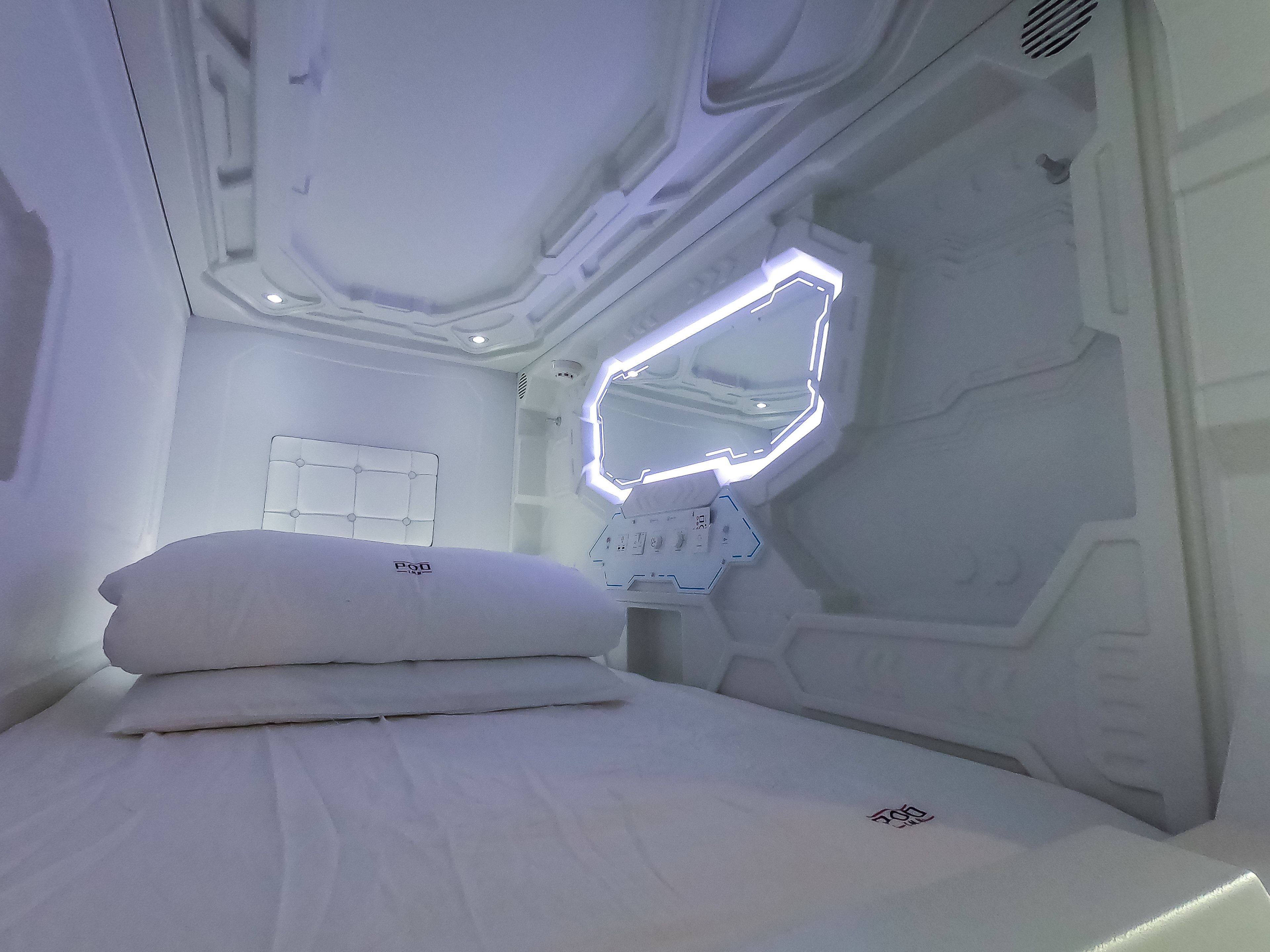 A modern, minimalist bedroom with a neatly made bed, a wall-mounted mirror, and a control panel.