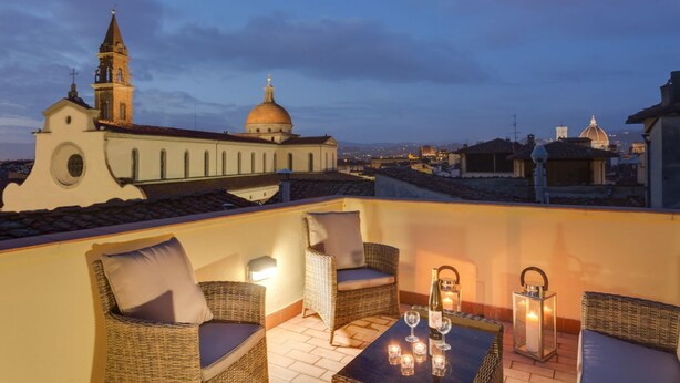 A rooftop terrace with wicker furniture, candles, and a view of historic buildings.
