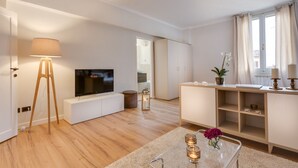 A modern living room with a flat-screen TV, a standing lamp, a wooden floor, a white cabinet, and a window with curtains.