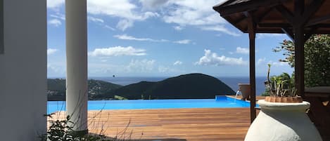 Una terraza de madera que da a una piscina con vistas al océano y a las colinas.