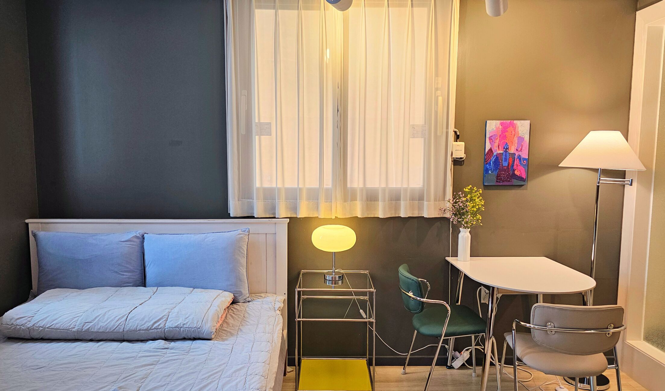 Een slaapkamer met een bed, een bureau, twee stoelen, een lamp, een vaas met bloemen en een schilderij aan de muur.