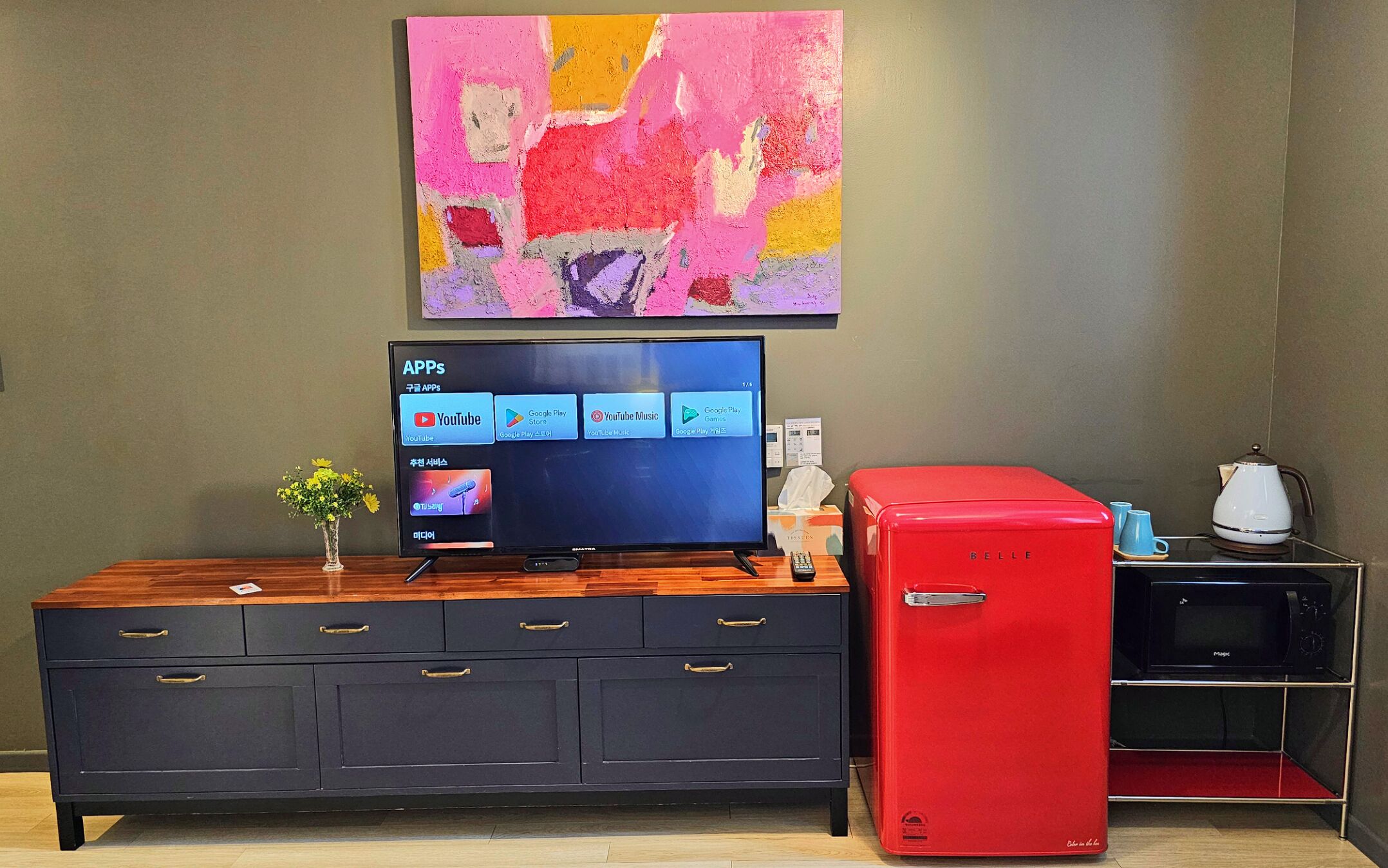 Una sala de estar moderna con un televisor de pantalla plana, un refrigerador rojo, un armario de madera y un cuadro abstracto colorido en la pared.