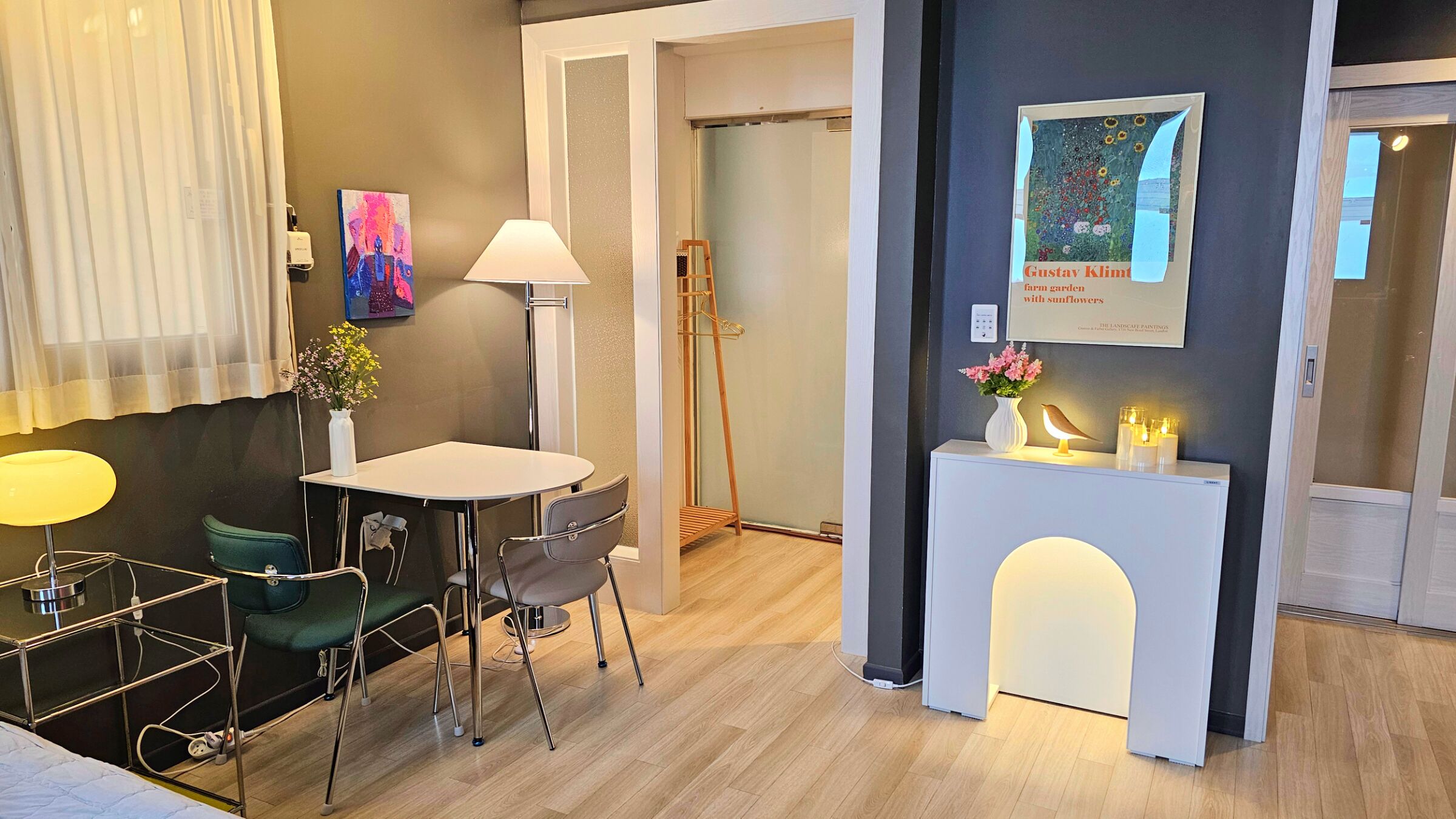 A modern hotel room with a dining area, a standing lamp, a painting, and a framed artwork on the wall.