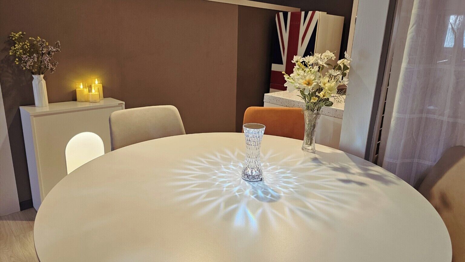 Uma mesa redonda com um centro de mesa de vidro transparente, um vaso com flores e velas sobre uma mesinha lateral branca.
