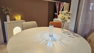 Uma mesa redonda com um centro de mesa de vidro transparente, um vaso com flores e velas sobre uma mesinha lateral branca.