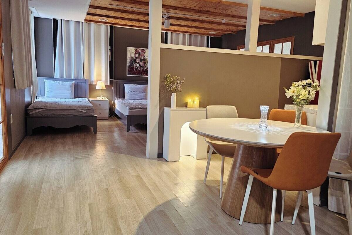 A modern hotel room with a wooden floor, a round dining table with chairs, a bed with a headboard, and a small side table with a lamp.