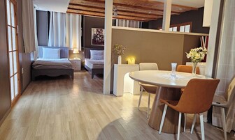 Una habitación de hotel moderna con suelo de madera, una mesa de comedor redonda con sillas, una cama con cabecero y una pequeña mesita de noche con lámpara.