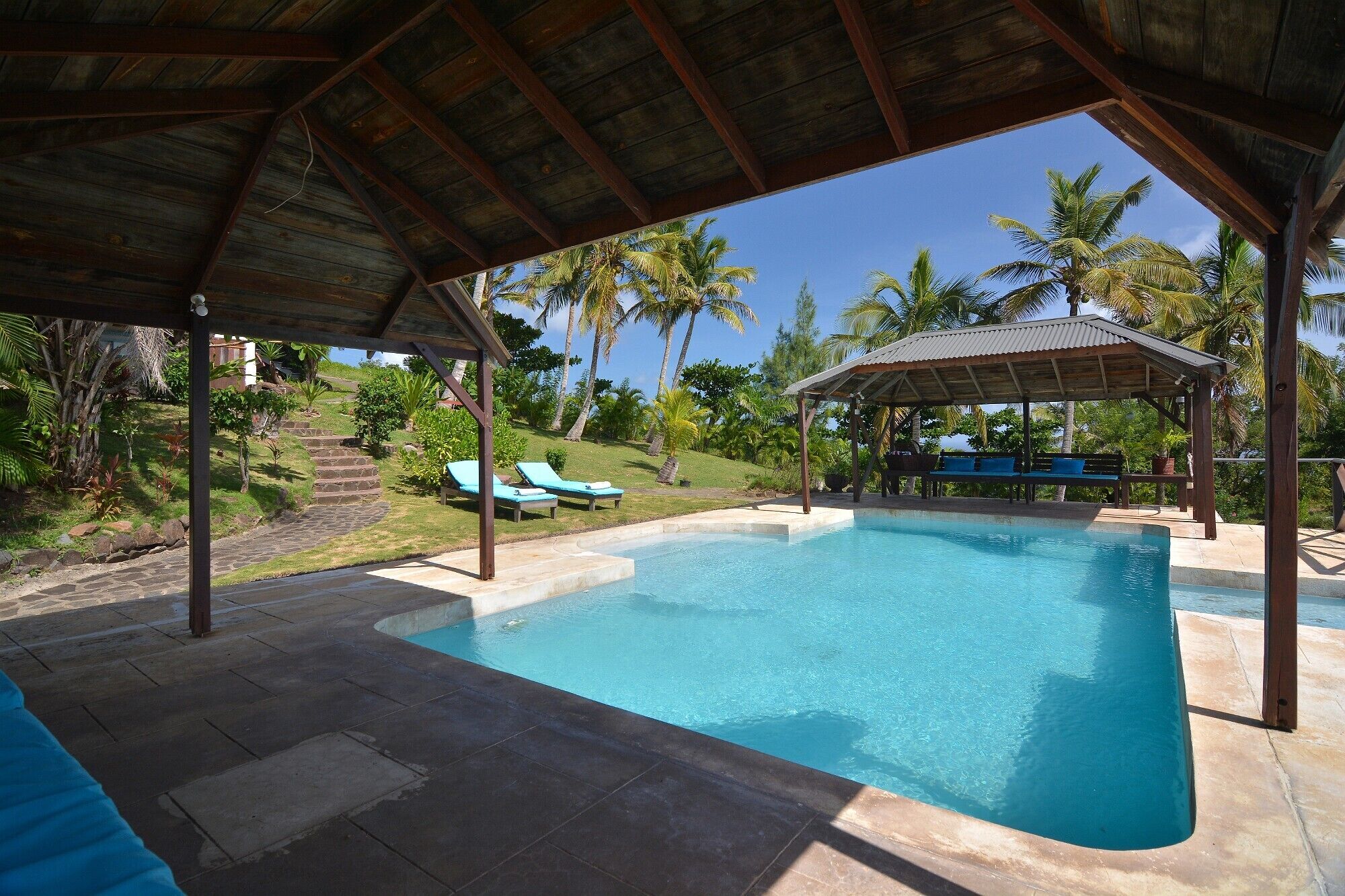 A pool area with a covered seating area, lounge chairs, and a view of palm trees and greenery.