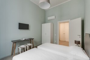 A bedroom with a bed, a desk, two chairs, a TV, a wardrobe, and an open door leading to another room.
