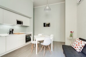 A modern kitchen with white cabinets, a microwave, and a coffee maker. There is a dining table with white chairs and a black sofa with a polka dot pillow.