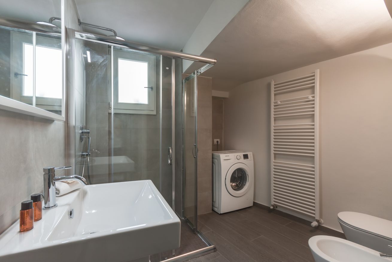 A modern bathroom with a glass-enclosed shower, a built-in sink, a washing machine, and a toilet.